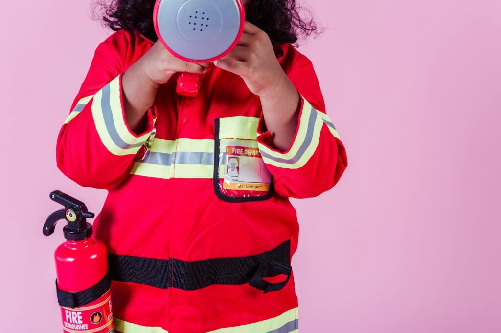 Crop unrecognizable little ethnic kid wearing fireman costume and fire extinguisher while using loudspeaker toy and standing on pink background