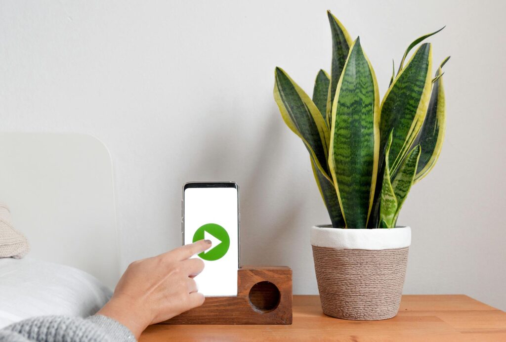 Crop unrecognizable female lying on comfy bed and pressing ply soft button on modern smartphone placed on creative wooden holder on bedside table