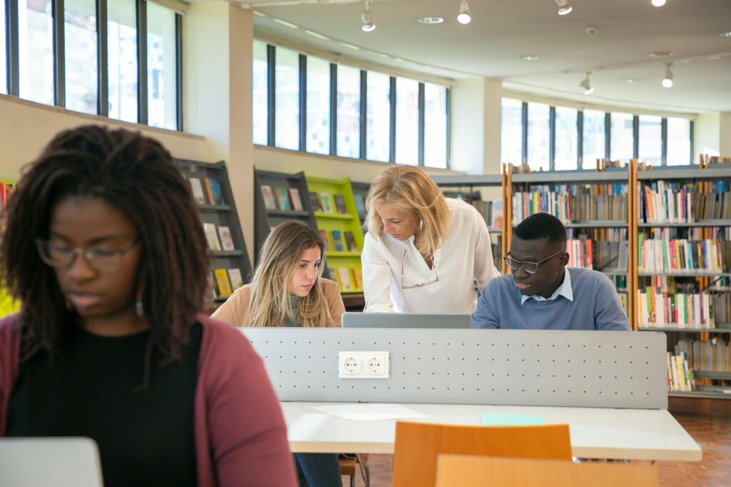 Elegant middle aged female library worker helping to young multiracial students to find information during work on laptop